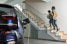 An electric car sitting in a specialized residential garage in the foreground, while a parent and child climb the stairs into their home in the background.